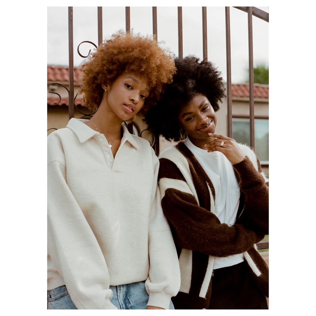 Two young African American women pose in front of a metal gate outdoors. One wears a beige sweater, the other a brown and white cardigan.