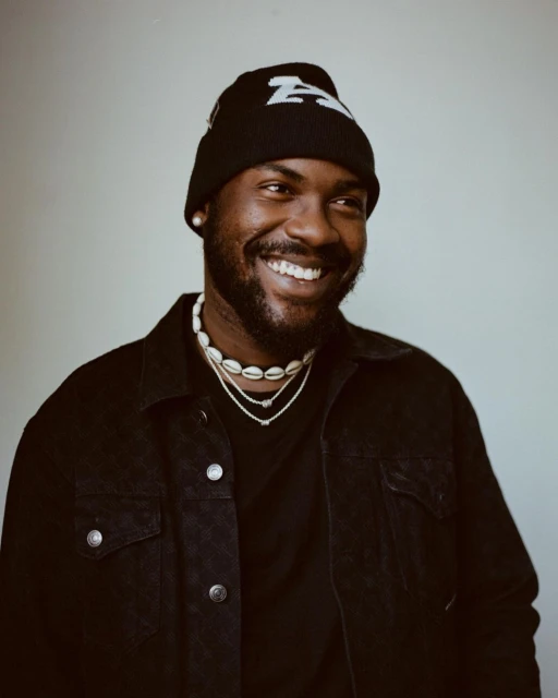 Close-up portrait of a smiling black man wearing a black beanie, necklaces, and a black jacket against a plain light gray background.