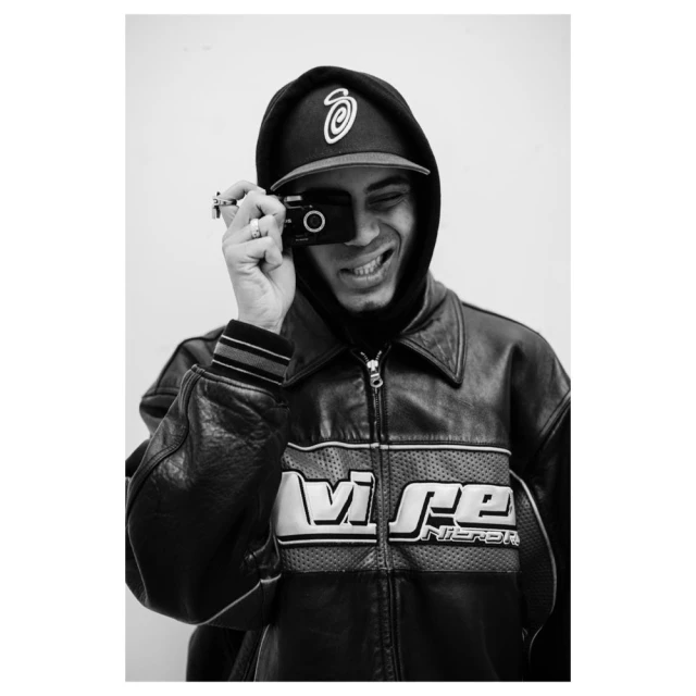 A person in a black hooded cap and leather jacket smiles while holding a camera up to their eye, set against a plain white backdrop.