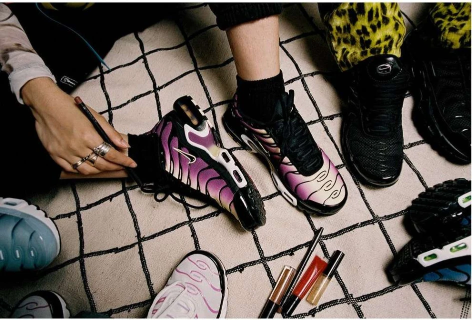 Close-up of several pairs of black and colorful Nike Air Max Plus sneakers, one being customized with makeup on a checkered rug indoors.