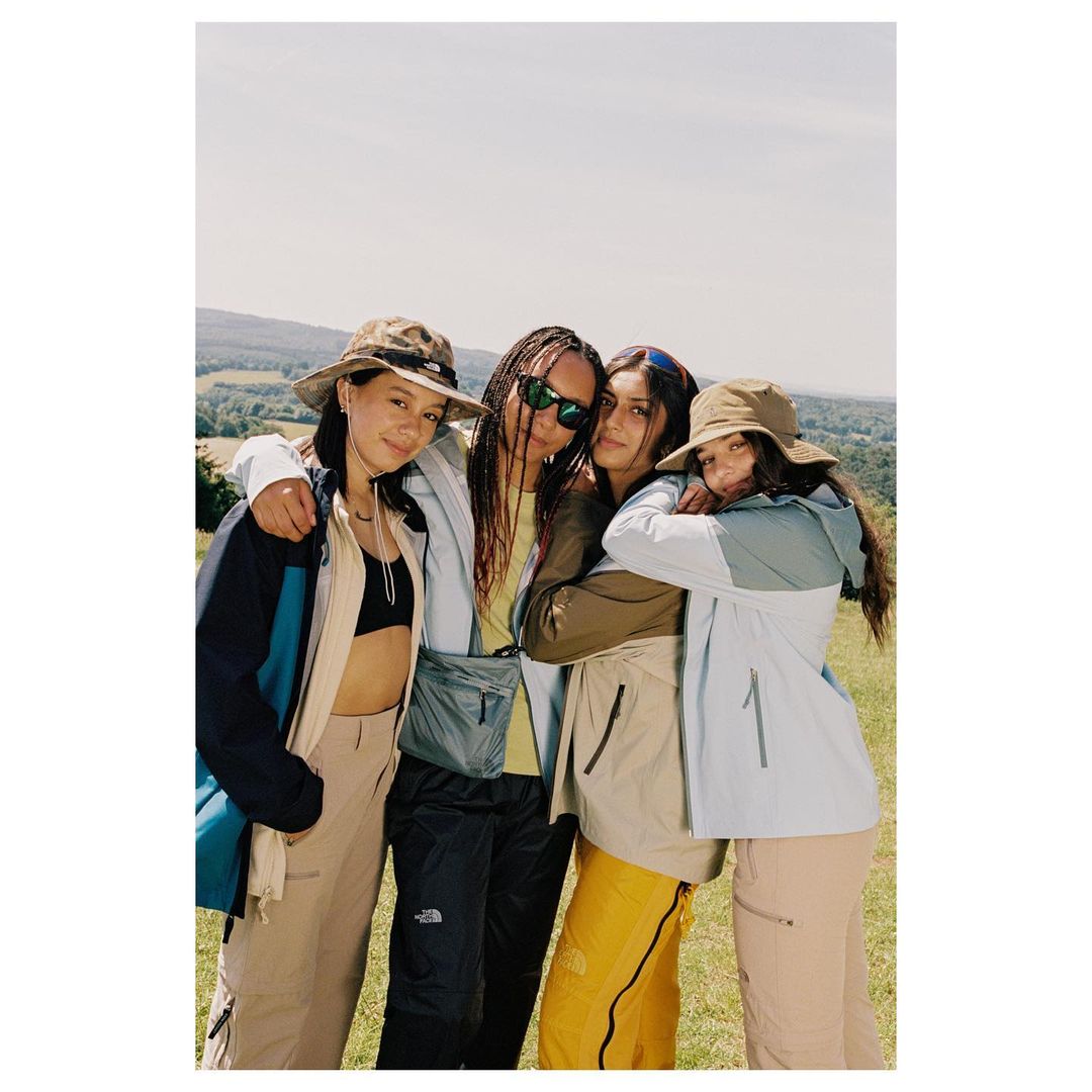 Four young women wearing outdoor apparel pose together on a grassy hillside with a lightly-hazy sky above and a forested area in background.