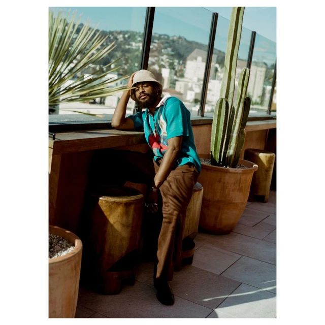 Man in turquoise polo and brown pants sitting on a wooden stool on an outdoor patio with cacti and a city view in the background.