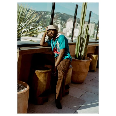 Man in turquoise polo and brown pants sitting on a wooden stool on an outdoor patio with cacti and a city view in the background.