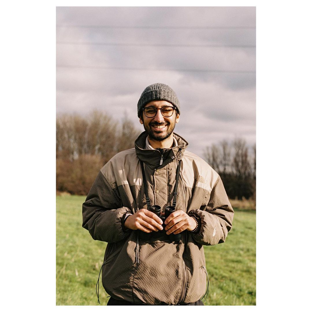 A smiling man with glasses and a beanie outdoors, holding binoculars and wearing a patterned jacket in a grassy field on a cloudy day.
