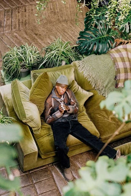 Person lounges on an olive green couch, surrounded by indoor plants, wearing a sheer jacket, beanie, jeans, and tan shoes, in a relaxed setting.