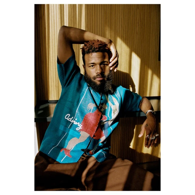 Stylish man with a beard, wearing a short-sleeved patterned shirt and brown pants, poses indoors with soft lighting.