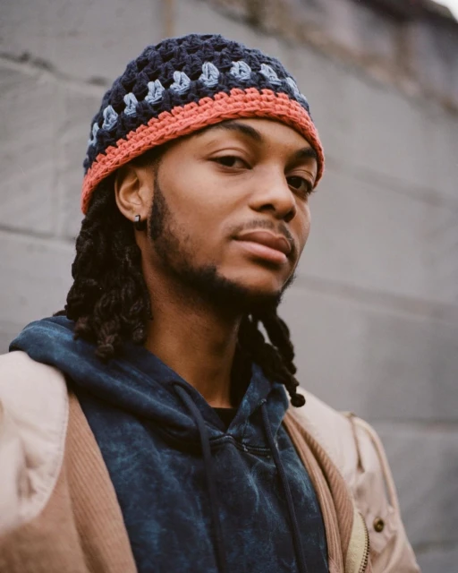 Portrait of a young African-American man wearing a crocheted hat and hooded sweatshirt, posing outdoors against a textured wall.