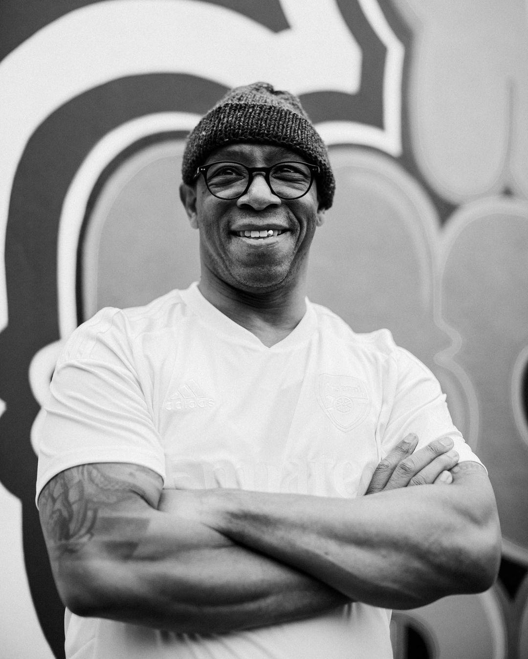 A smiling adult male with glasses and a beanie stands indoors with arms crossed against a graffiti wall in a medium shot portrait.