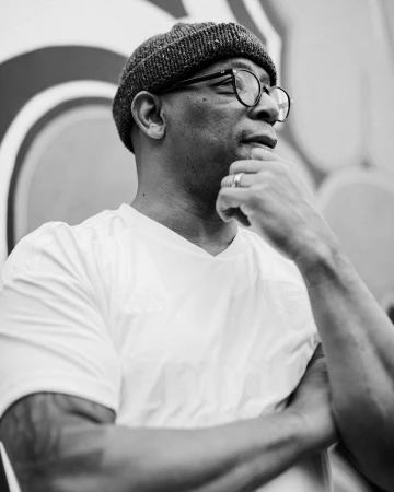 Black and white portrait of a black man wearing glasses, a beanie, and a white t-shirt, photographed by Vicky Grout in London.