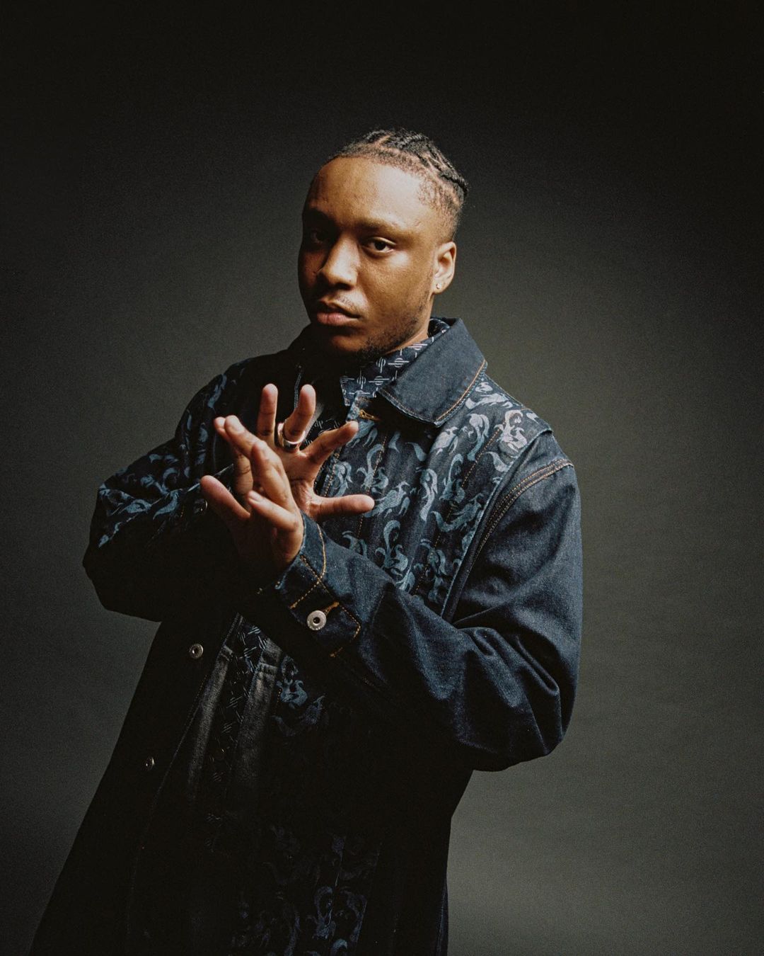 Portrait of a young adult male with braided hair, wearing a patterned denim jacket in a studio setting against a gray backdrop.