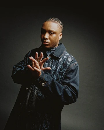 Portrait of a young adult male with braided hair, wearing a patterned denim jacket in a studio setting against a gray backdrop.