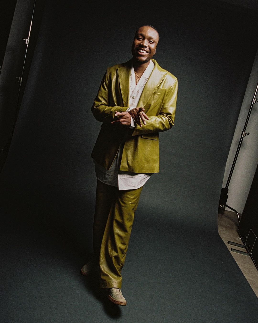 A smiling Black man poses in a mustard-color, textured suit with a white shirt, against a dark grey backdrop in a studio with lighting equipment.