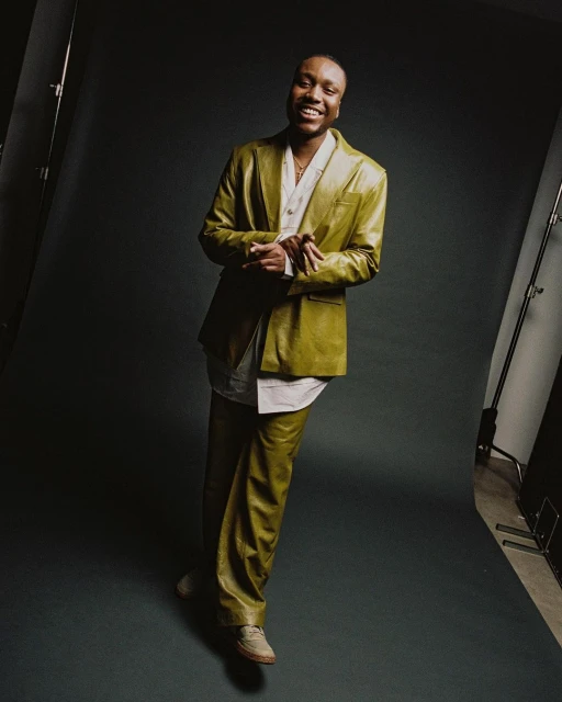 A smiling Black man poses in a mustard-color, textured suit with a white shirt, against a dark grey backdrop in a studio with lighting equipment.