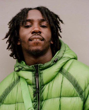 Portrait of a young adult male with dreadlocks wearing a bright green puffer jacket and standing indoors against a light grey wall.