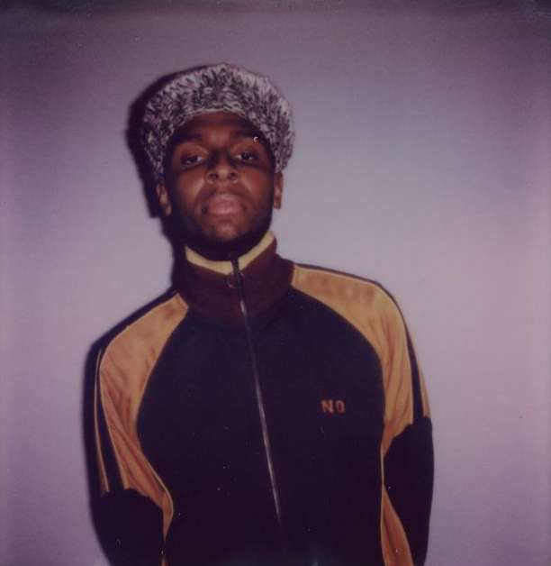 Portrait of a Black adult male in a track jacket and textured hat against a neutral gray backdrop, lit by soft, diffused studio lighting.