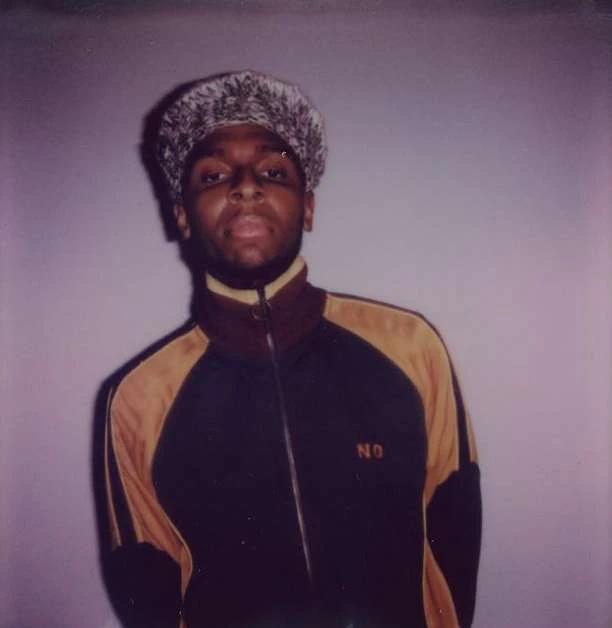 Portrait of a Black adult male in a track jacket and textured hat against a neutral gray backdrop, lit by soft, diffused studio lighting.
