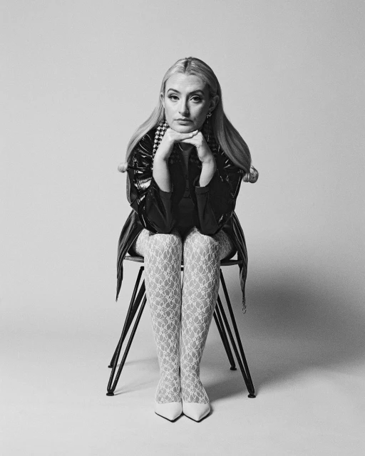 Blonde woman in a black, shiny jacket and patterned white tights sits on a black chair in a minimalist, black and white portrait.