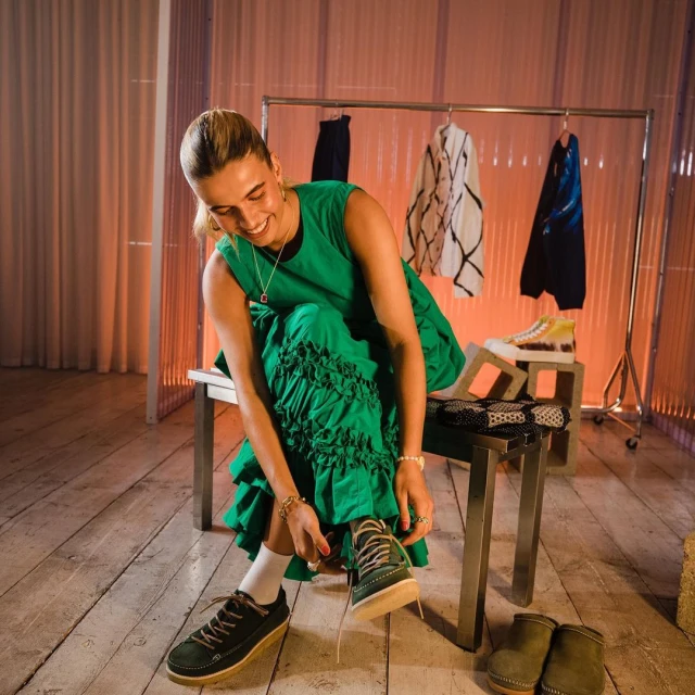 A smiling woman in a green dress sits on a stool and ties her shoe in a studio setting, with clothing rack and shoes visible in background.