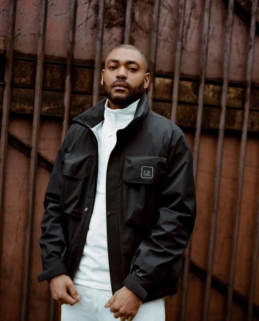 Portrait of a black man wearing a black zip jacket and white turtleneck against a rustic metal gate background.