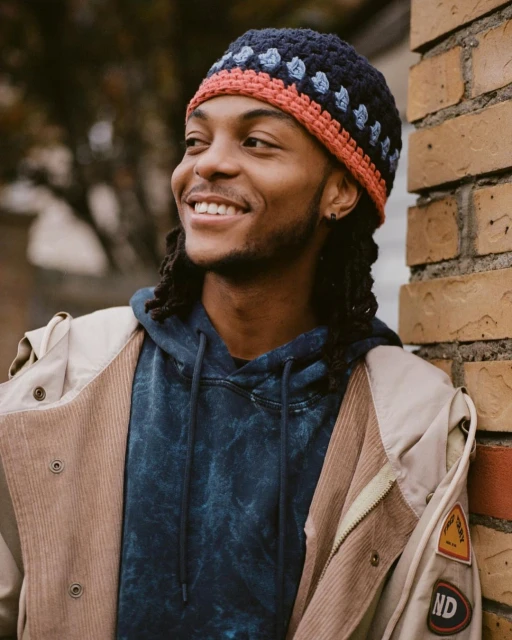 Young man wearing a colorful crochet beanie and jacket smiles while leaning against a brick wall outdoors in an eye-level shot.
