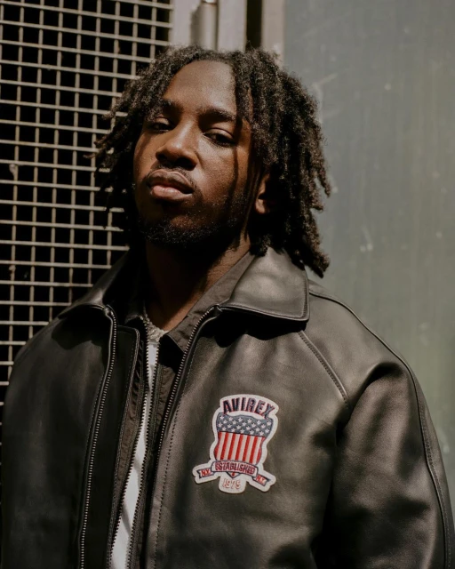 Portrait of a Black male model wearing a leather jacket with a patch, shot indoors against a metal grid backdrop.