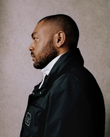 Profile portrait of a Black man with short dark hair and beard wearing a black jacket over a white collared shirt against a textured wall