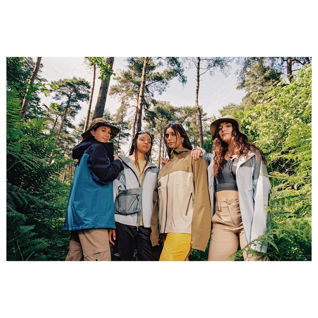 A group of four young adult women are standing close together in a lush forest, all wearing outdoor-style jackets and hats in natural light.