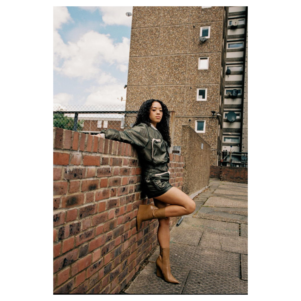 Woman in olive green jacket and shorts leans against brick wall in urban setting, wearing heeled boots and posing for fashion photo.