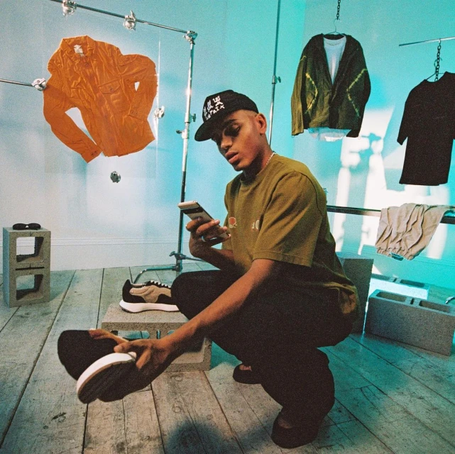 Person crouching indoors, using a smartphone, surrounded by fashion clothing racks with shirts, and cinder blocks on the wooden floor.