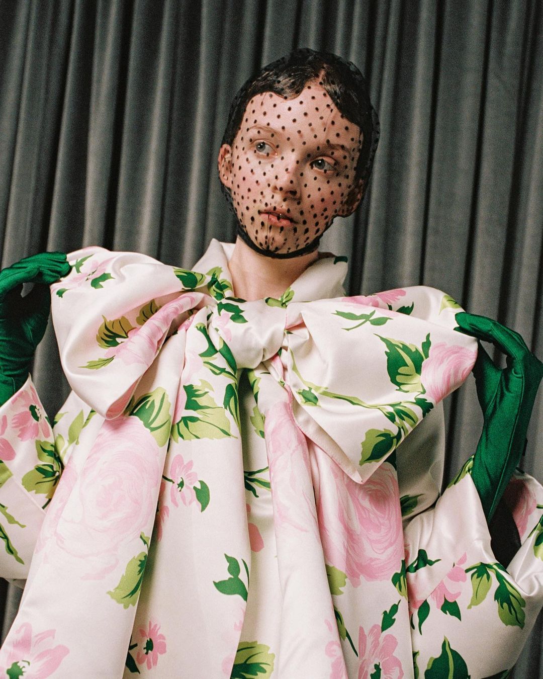 Woman poses in studio with oversized floral dress, green gloves and dotted veil. Fashion shot features unique couture and design elements.