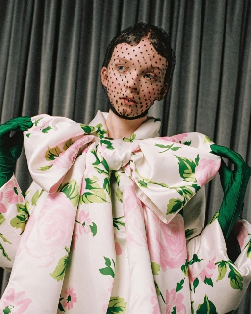 Woman poses in studio with oversized floral dress, green gloves and dotted veil. Fashion shot features unique couture and design elements.