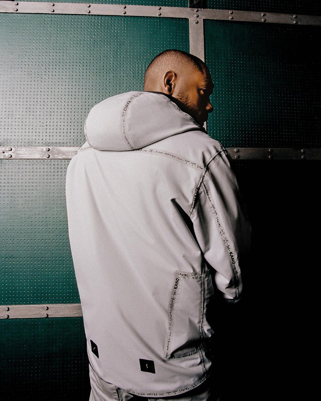Black man wearing a light grey hooded anorak with printed trim, captured from behind against a textured green industrial metal wall.