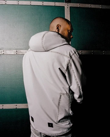 Black man wearing a light grey hooded anorak with printed trim, captured from behind against a textured green industrial metal wall.