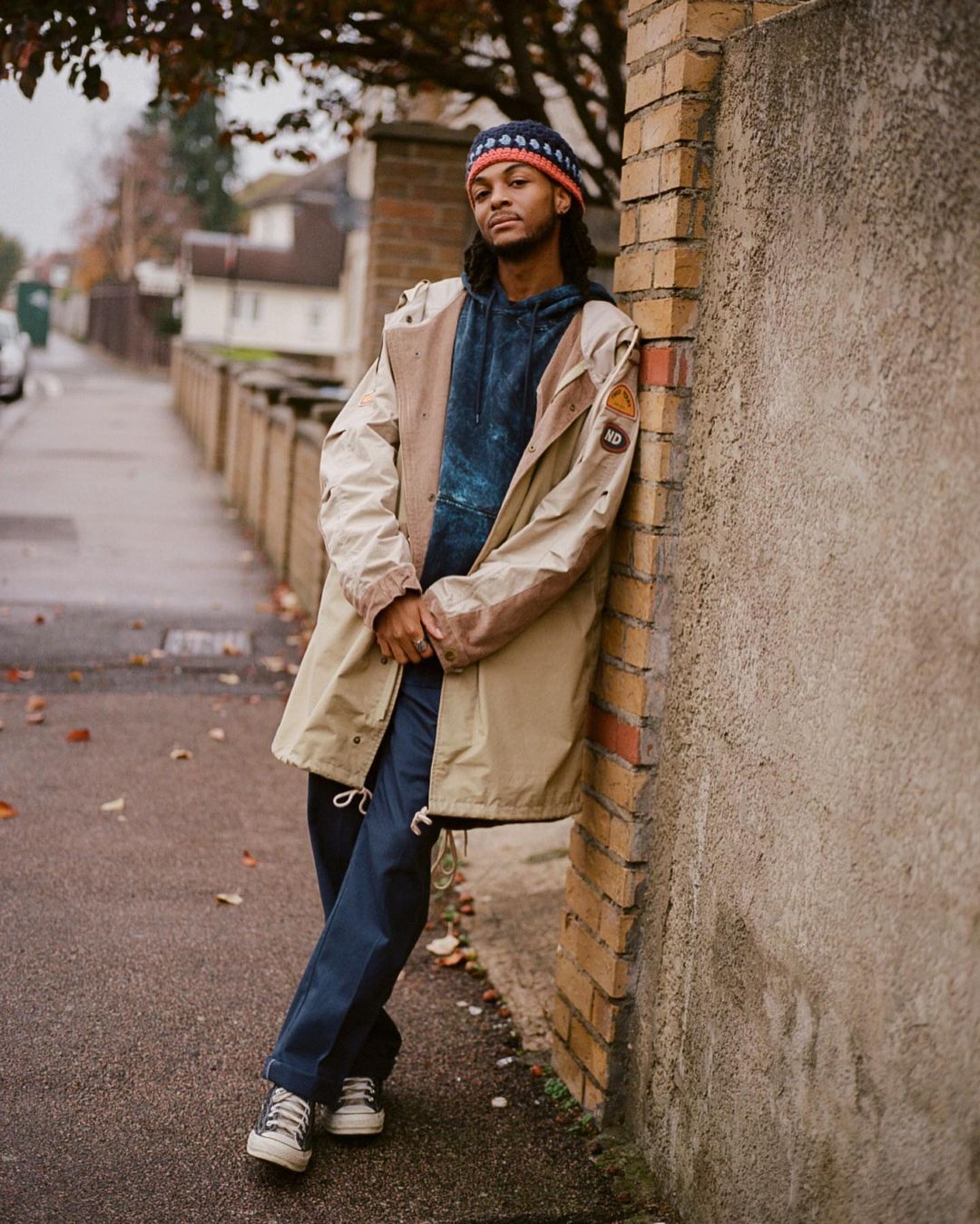 Adult male in jacket and beanie leaning against a brick wall on a sidewalk with trees and houses in the background on an overcast day.
