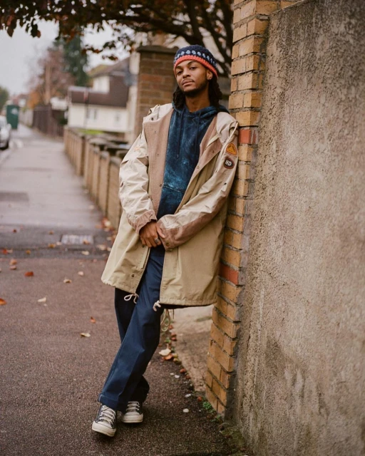 Adult male in jacket and beanie leaning against a brick wall on a sidewalk with trees and houses in the background on an overcast day.