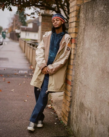 Adult male in jacket and beanie leaning against a brick wall on a sidewalk with trees and houses in the background on an overcast day.
