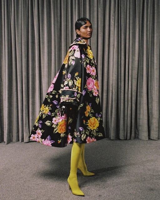 Woman in floral coat and yellow stockings poses against grey drapes in studio. Fashion editorial shot, eye-level angle, medium view.