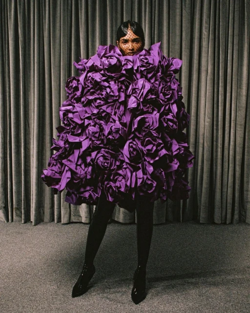 An adult female model poses in a high-fashion setting, wearing a large purple rose-like dress, tights, and heels against a curtain backdrop.