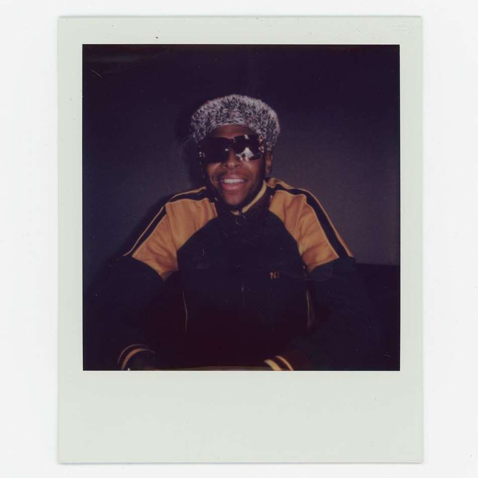 Person smiling in dark jacket, light fuzzy hat, and sunglasses, sitting indoors against a grey backdrop. Polaroids frame.