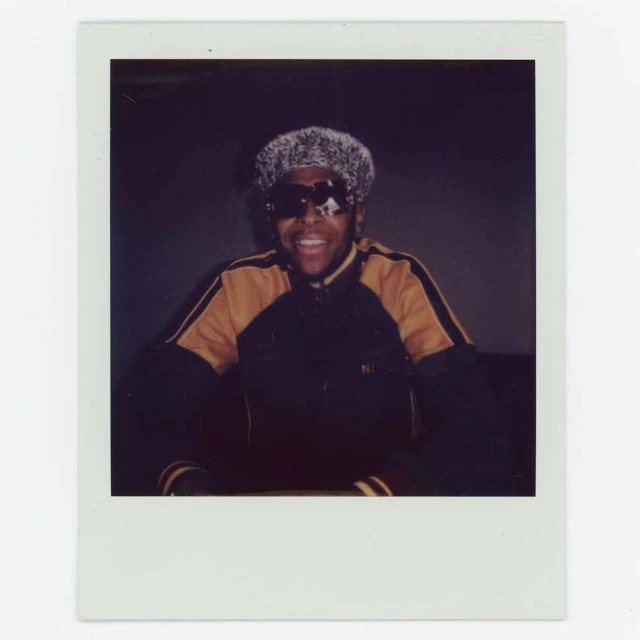 Person smiling in dark jacket, light fuzzy hat, and sunglasses, sitting indoors against a grey backdrop. Polaroids frame.