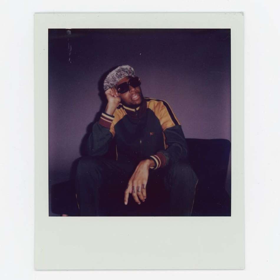 Polaroid photo of an adult male sitting, wearing dark sunglasses and a patterned hat, in a dark green and yellow jacket.