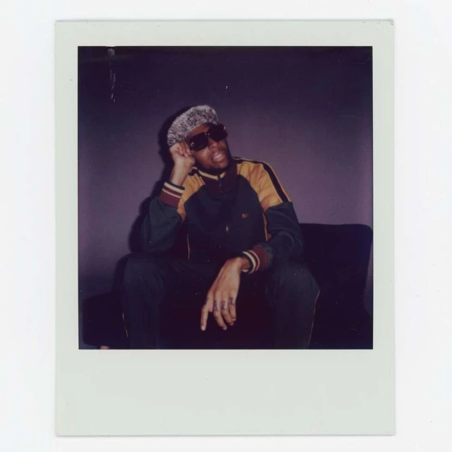 Polaroid photo of an adult male sitting, wearing dark sunglasses and a patterned hat, in a dark green and yellow jacket.