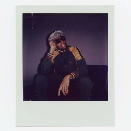 Polaroid photo of an adult male sitting, wearing dark sunglasses and a patterned hat, in a dark green and yellow jacket.