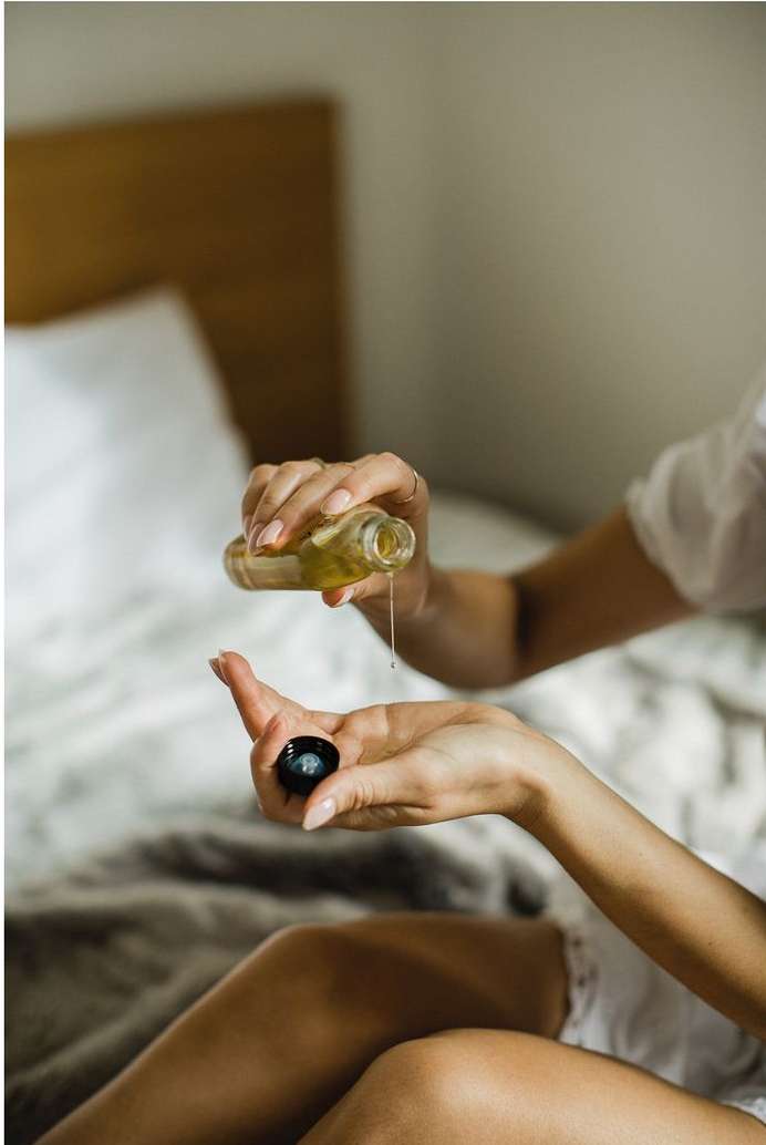 Close-up of a woman pouring oil from a clear glass bottle into the palm of her hand, set in a softly lit room with a neutral backdrop.