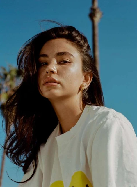 Close-up portrait of a young adult female with long dark hair and freckles, wearing a white t-shirt against a bright blue sky.
