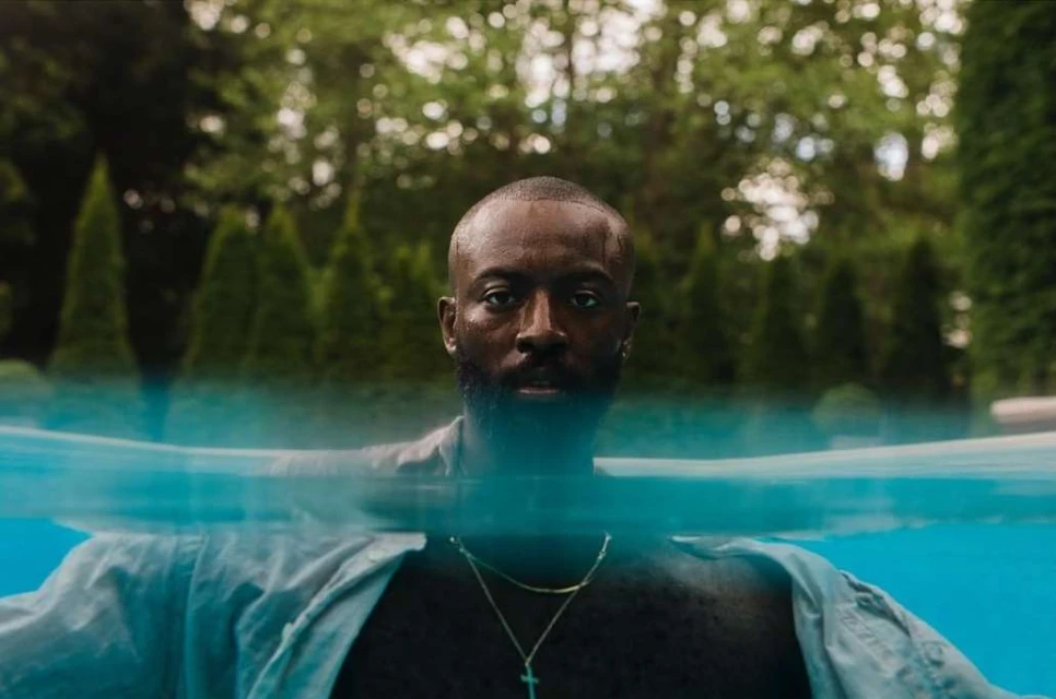 A man with a beard swims in a pool outdoors, partially submerged in water. Green trees are visible in the background.