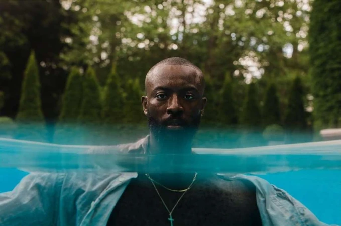 A man with a beard swims in a pool outdoors, partially submerged in water. Green trees are visible in the background.