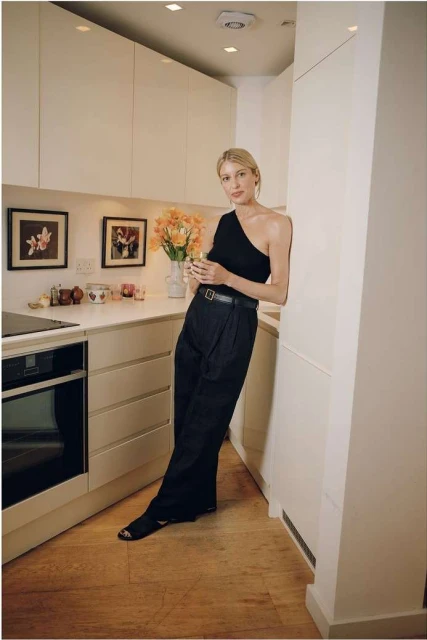 Adult female in one-shoulder top and wide-leg pants, leaning against a wall in a light and airy kitchen setting with modern appliances.