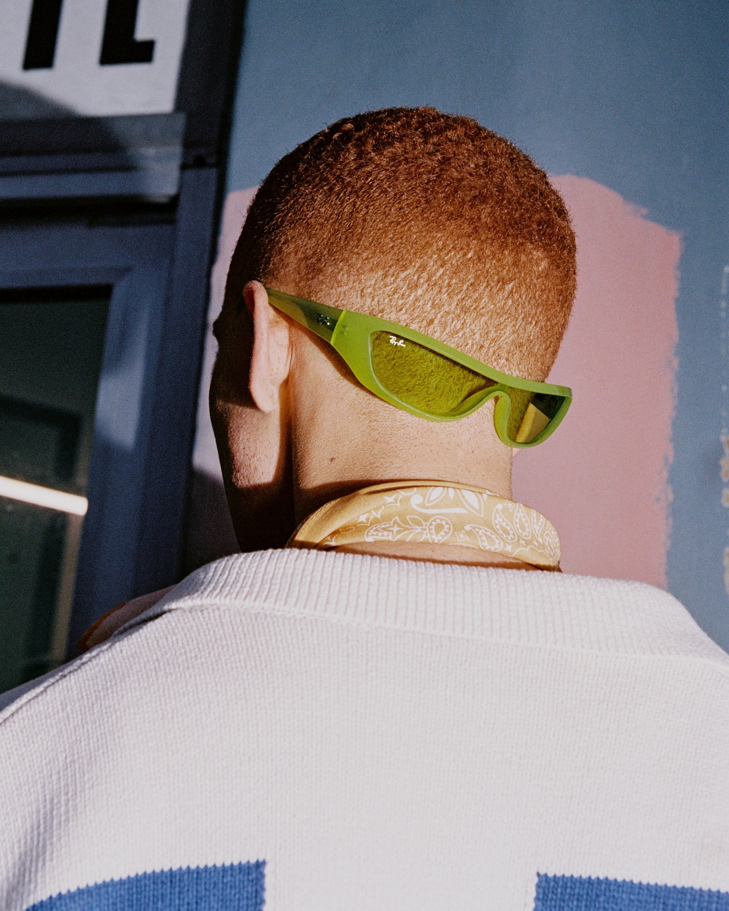Rear view of a person with a buzz cut, wearing sunglasses and a patterned bandana, facing away from the camera with a blue background.
