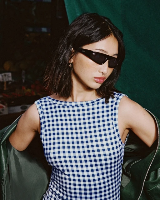 Brunette woman poses in blue-and-white checkered top and black sunglasses against a dark background with green fabric and fruit.